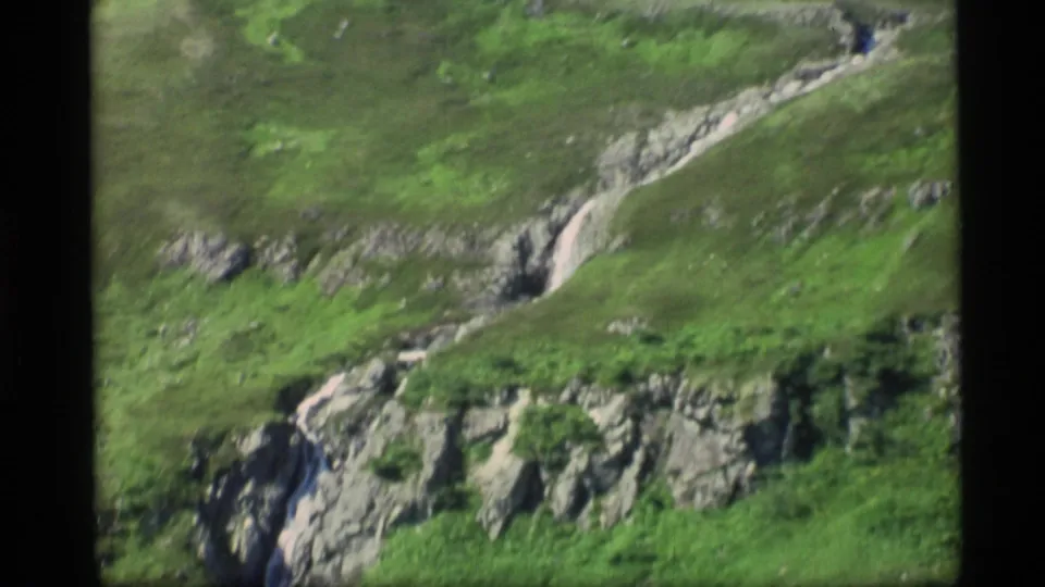 1977 Alaska 8mm frame of gray rock formations and bright vegetation around a thin stream on a steep slope.