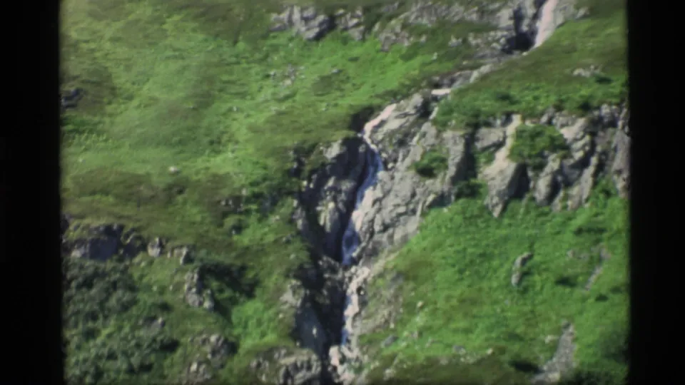 1977 Alaska 8mm frame showing a rugged ravine wall, scattered vegetation, and a narrow watercourse through broken rock.