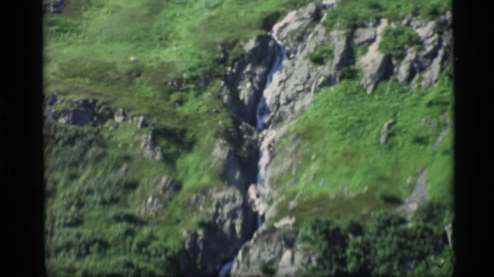 1977 Alaska 8mm frame of a narrow stream dropping through rocky terrain bordered by green hillside vegetation.