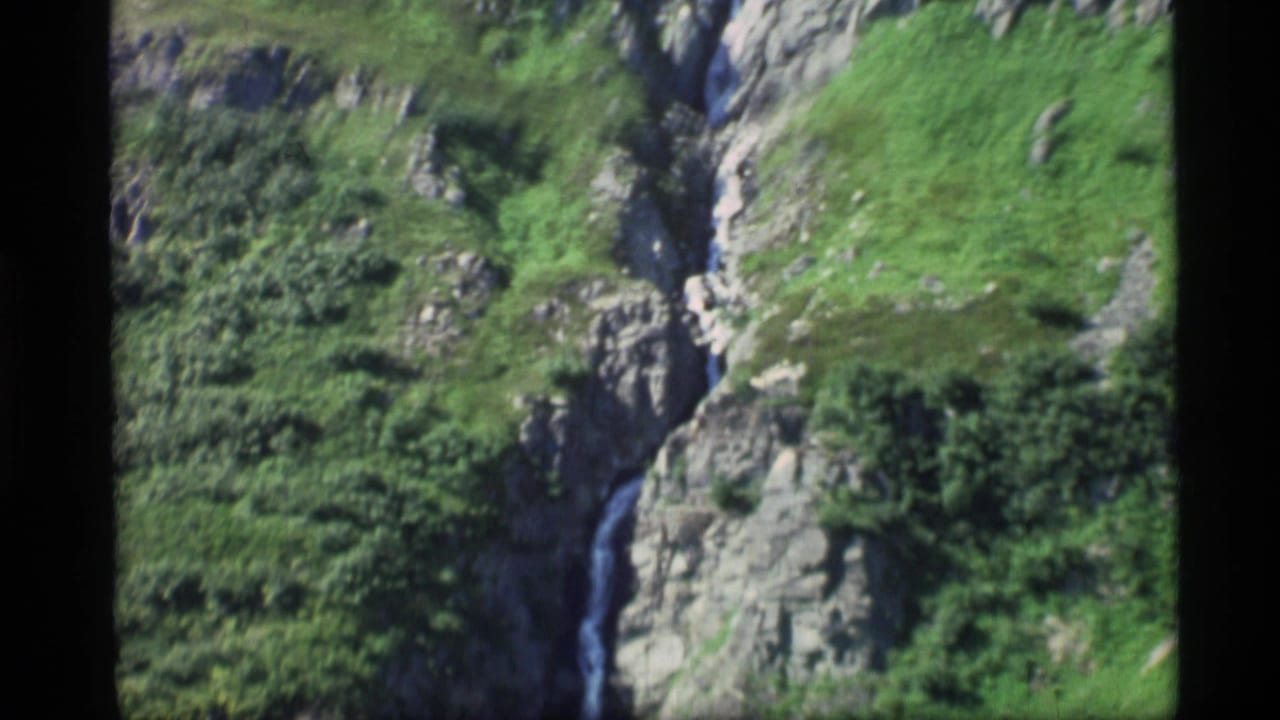 1977 Alaska 8mm frame of a narrow stream running down gray rocks amid green hillside vegetation.