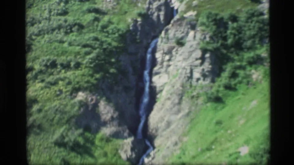 A thin Alaska mountain waterfall appears between gray rocks and dense green vegetation in this 1977 8mm frame.