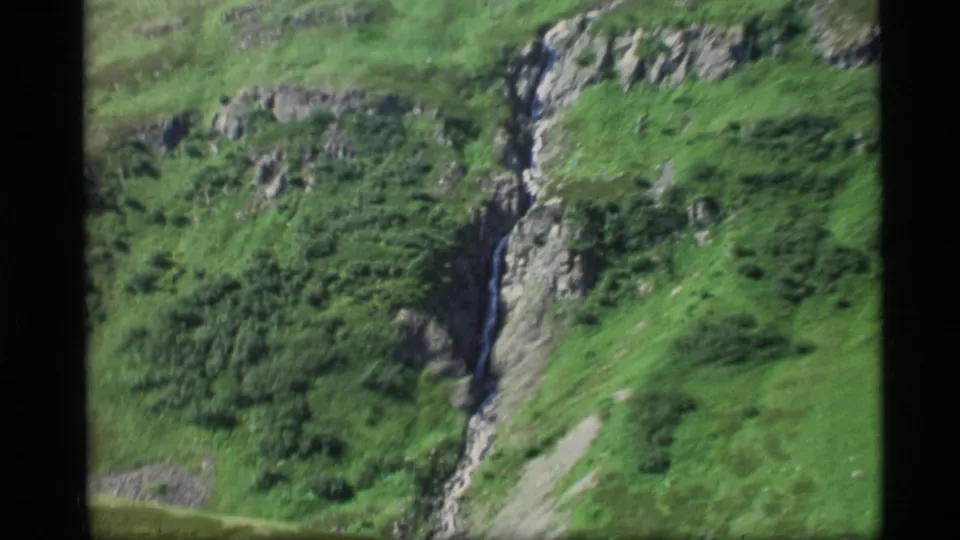 A narrow waterfall cuts through a gray rock face and bright green vegetation in 1977 Alaska home-movie footage.