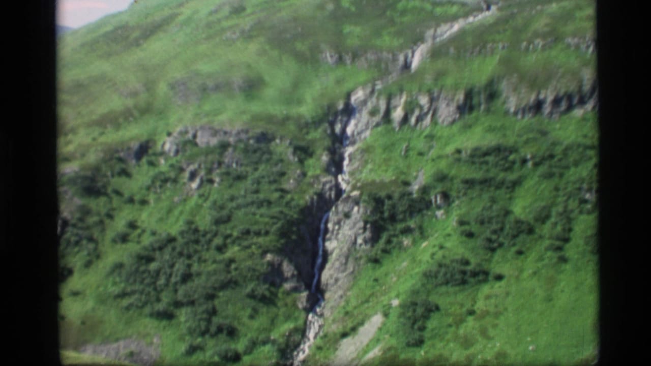 1977 Alaska 8mm frame showing a green mountain canyon, exposed rock, and a thin waterfall cutting down the hillside.