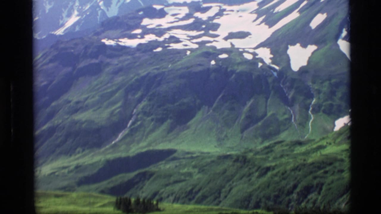 1977 Alaska 8mm frame showing a wide alpine valley, green slopes, snow patches, and distant mountain ridges.