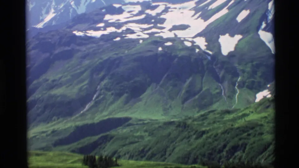 1977 Alaska 8mm frame showing a green alpine valley beneath snow-patched slopes, with a dark basin and small conifer stand below.