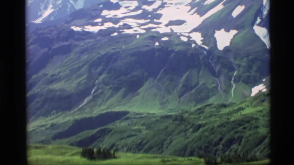 1977 Alaska 8mm frame showing snow-patched mountains above a green valley, with dark conifers near the lower edge.