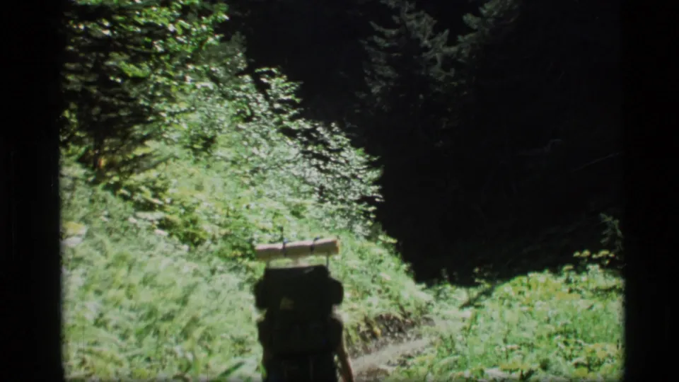 A backpacked hiker walks away along a narrow green forest trail in the 1977 Alaska 8mm footage.