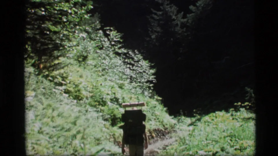 A backpacked hiker walks away along a sunlit forest path in 1977 Alaska home-movie footage.