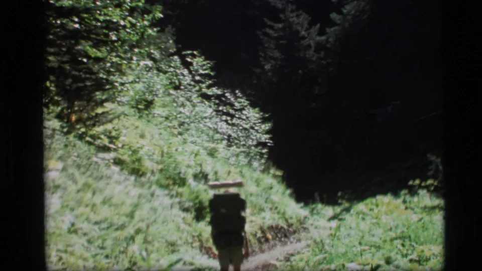 A distant hiker with a large pack follows a narrow green trail through Alaska forest footage from 1977.