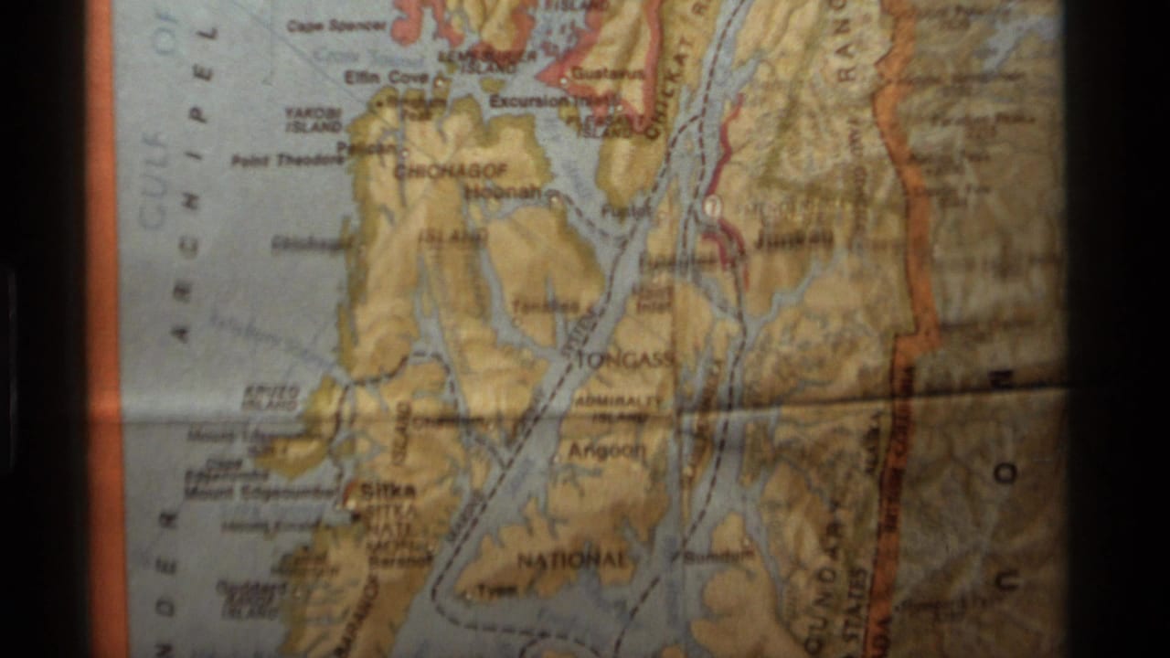 1977 8mm frame of a printed Alaska map section with coastline, islands, waterways, and border-area labels visible.