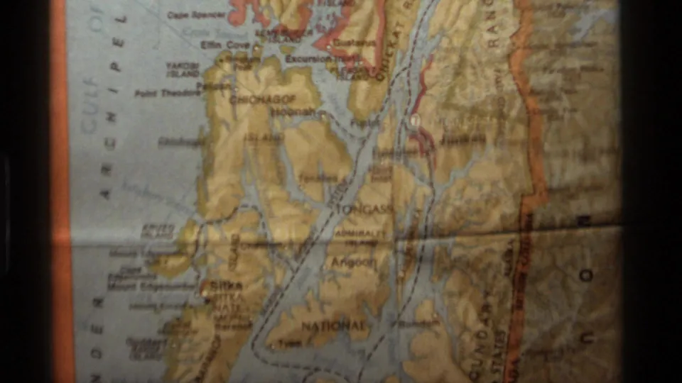 1977 8mm frame of a printed Alaska map section with coastline, islands, waterways, and border-area labels visible.