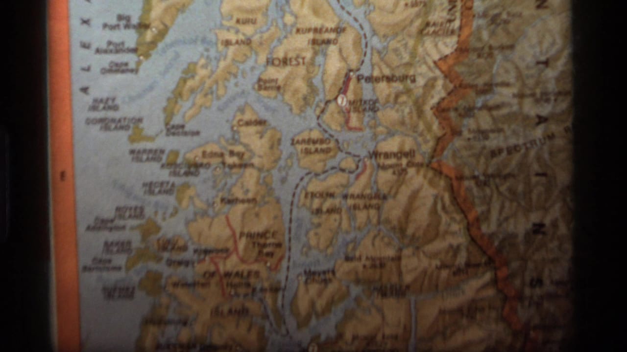 1977 8mm frame showing a close-up of an Alaska relief-style map with Southeast Alaska labels, shaded terrain, and orange map borders.