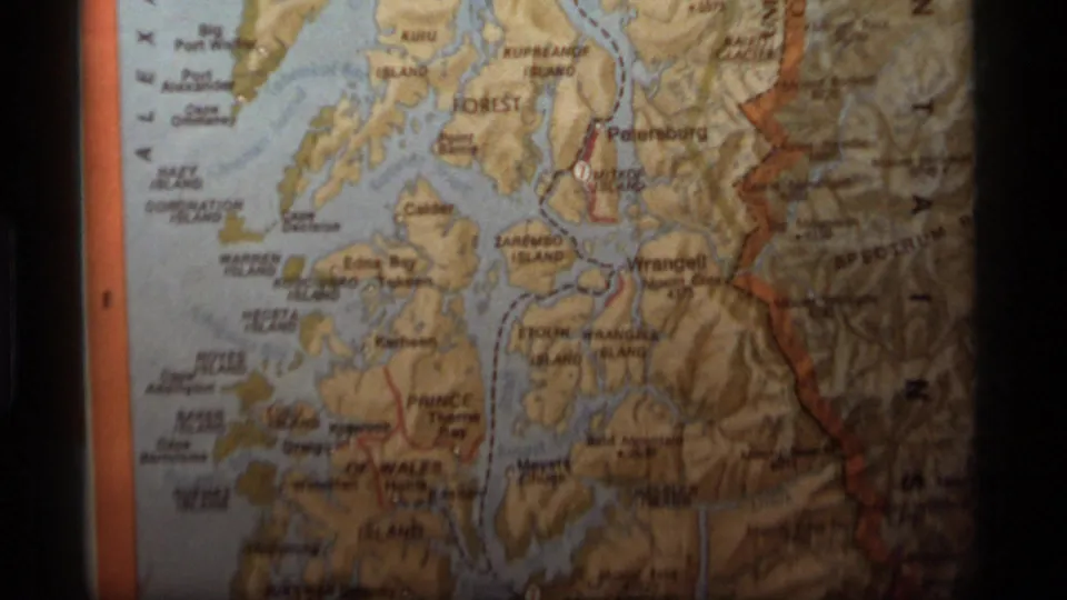 1977 8mm frame showing a close-up of an Alaska relief-style map with Southeast Alaska labels, shaded terrain, and orange map borders.