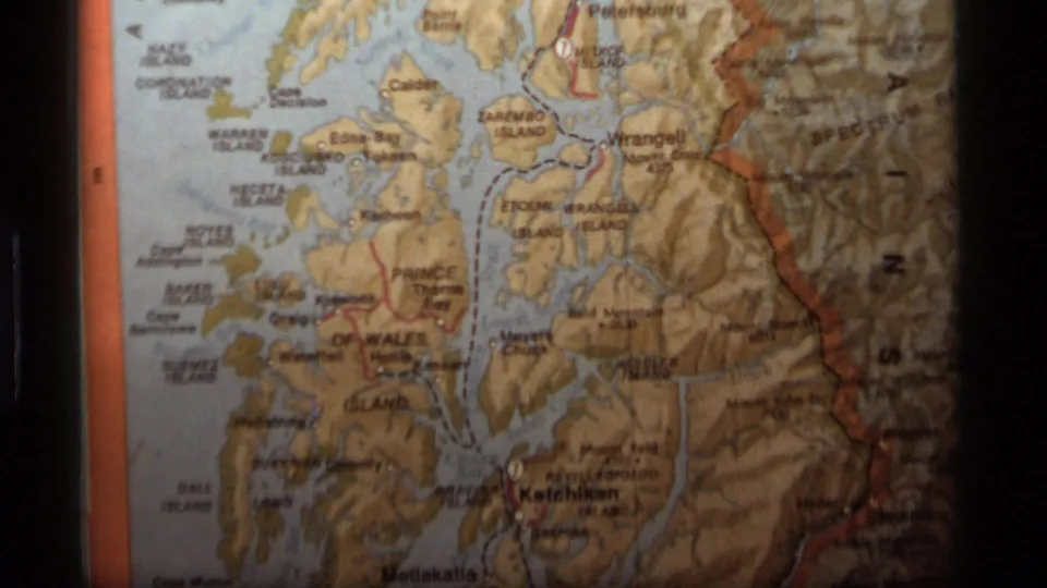 1977 home-movie frame of a printed Alaska relief map, showing lower coastal islands, Ketchikan-area text, and visible 8mm grain.
