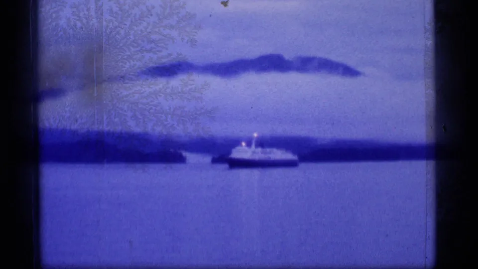 Blue-tinted 8mm Alaska travel frame showing a small boat on calm water below distant dark mountains.