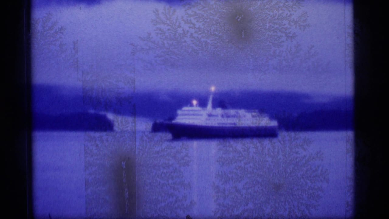 1977 Alaska 8mm frame showing a passenger ship moving across calm water with shoreline and misty mountains in the distance.