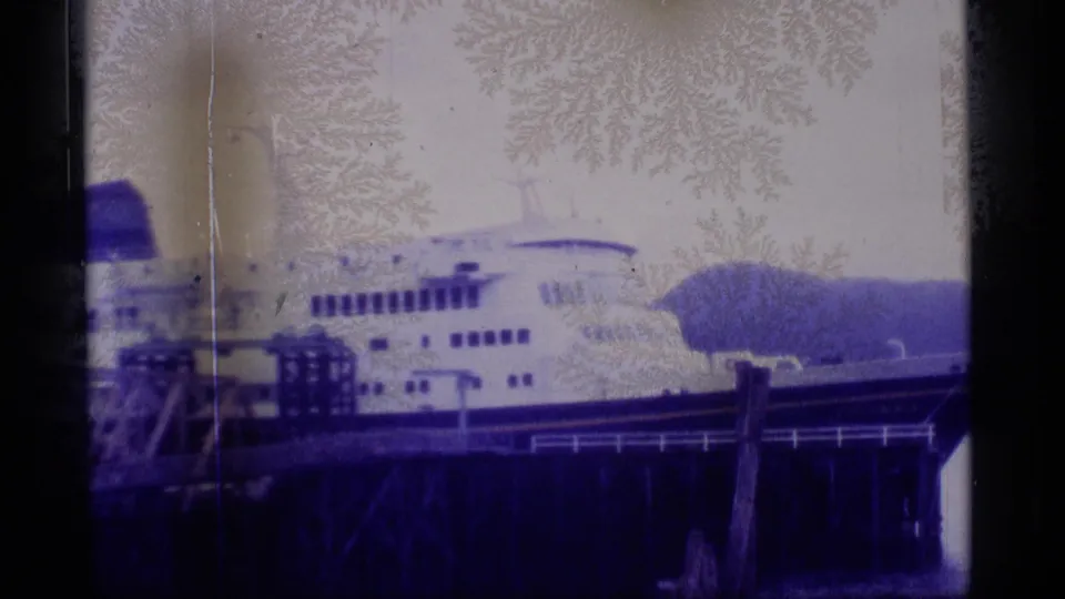 1977 Alaska frame showing the passenger ship at or near the dock, with railings, pilings, water, and shoreline visible.
