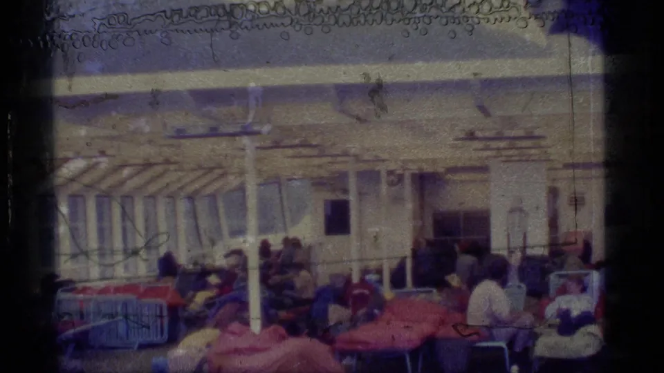 Passengers relax on a cruise ship deck in 1977 Alaska footage, with cots, chairs, posts, and awning structure visible.