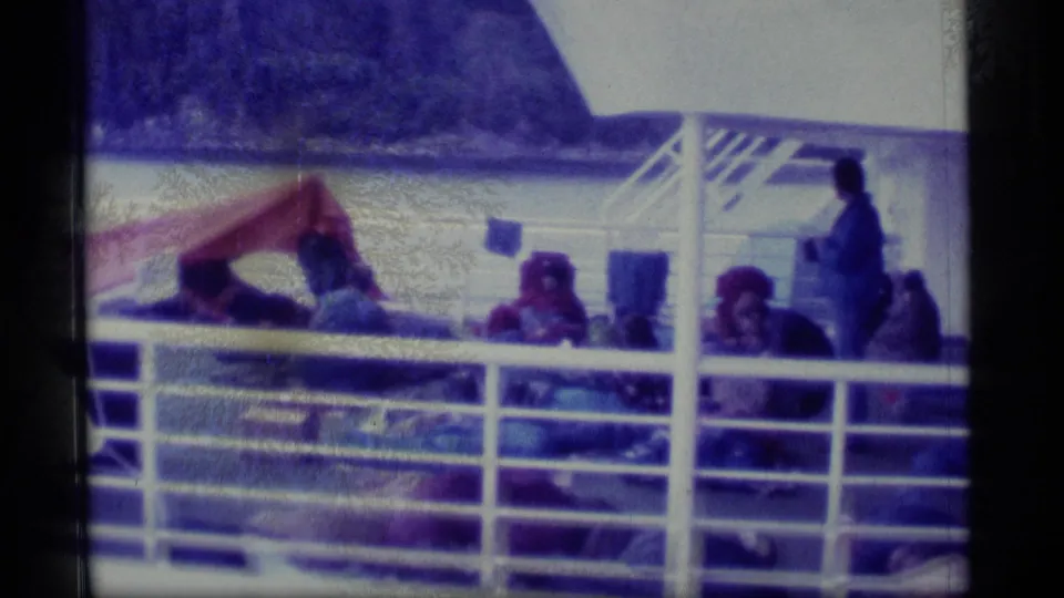 Passengers on an Alaska cruise deck sit behind white railings, with a mountainous shoreline visible in the background.