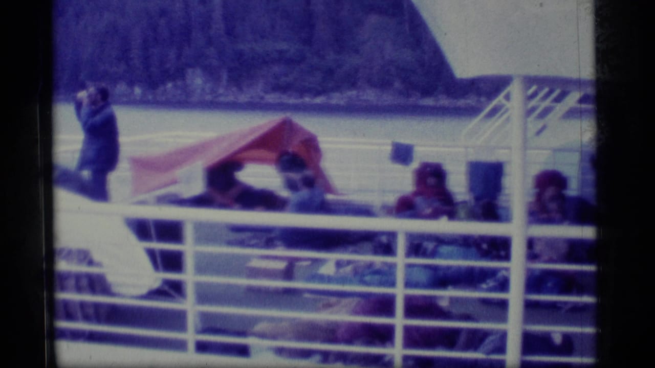 1977 Alaska home-movie frame showing travelers seated under a red tarp awning on a boat deck with water and mountains beyond.