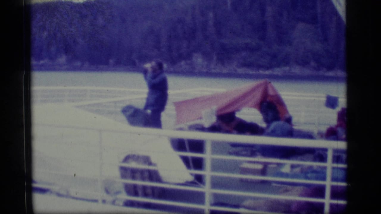 A standing passenger faces the water near a red tarp awning on a boat deck.