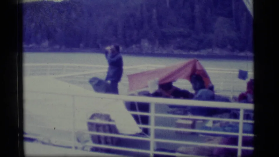 1977 Alaska boat-deck view with a standing traveler near seated passengers under a red tarp awning, water, and wooded mountains.