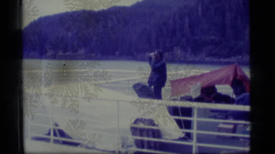 Soft 8mm frame from 1977 Alaska showing passengers on a boat deck beneath a red tarp awning with water and forested hills behind.