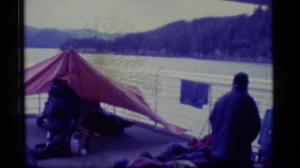 1977 Alaska home-movie frame with a rear-facing passenger near a red tarp awning, deck railings, calm water, and mountain shoreline.