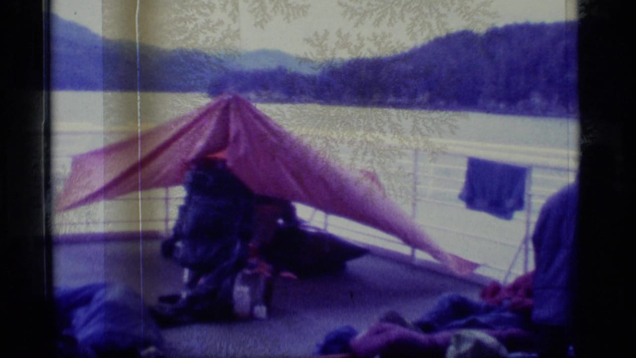 Travelers and gear on a boat deck beneath a red tarp with lake and mountains beyond.