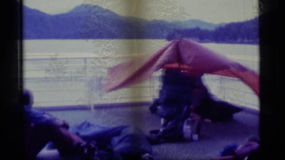 1977 Alaska travel footage frame with a red tarp over deck gear, a partially visible passenger, boat railing, lake water, and forested mountains.