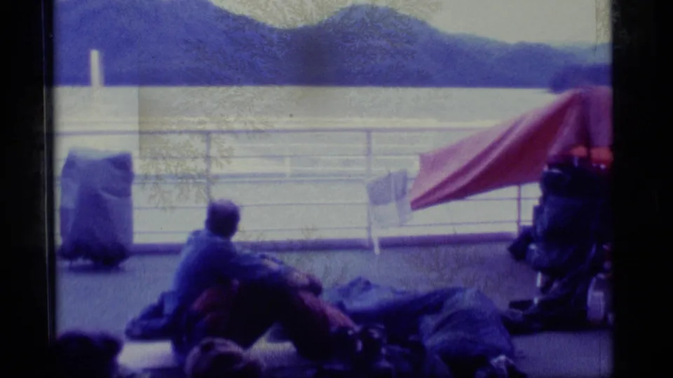 A 1977 Alaska home-movie frame shows a traveler seated among bags on an open boat deck, with a red tarp and mountain lake view behind.