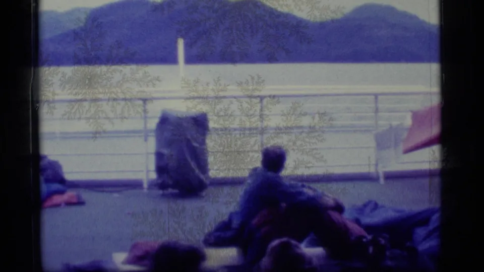 1977 Alaska 8mm frame of a seated traveler and covered gear on a boat deck, looking out toward a broad lake and dark mountain ridges.
