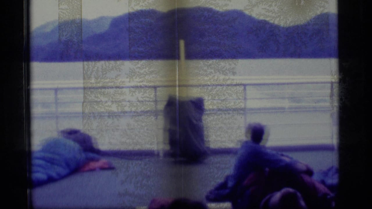 1977 Alaska 8mm frame showing passengers resting on an open ship deck as wooded mountains and still water pass beyond the rail.