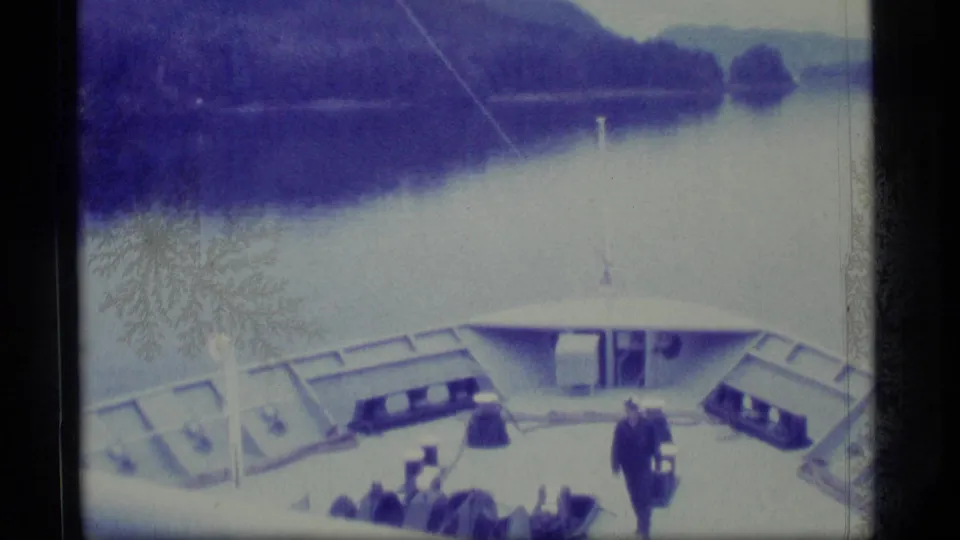 1977 Alaska home-movie frame looking over a ship bow toward calm water, shoreline, and forested mountain slopes.