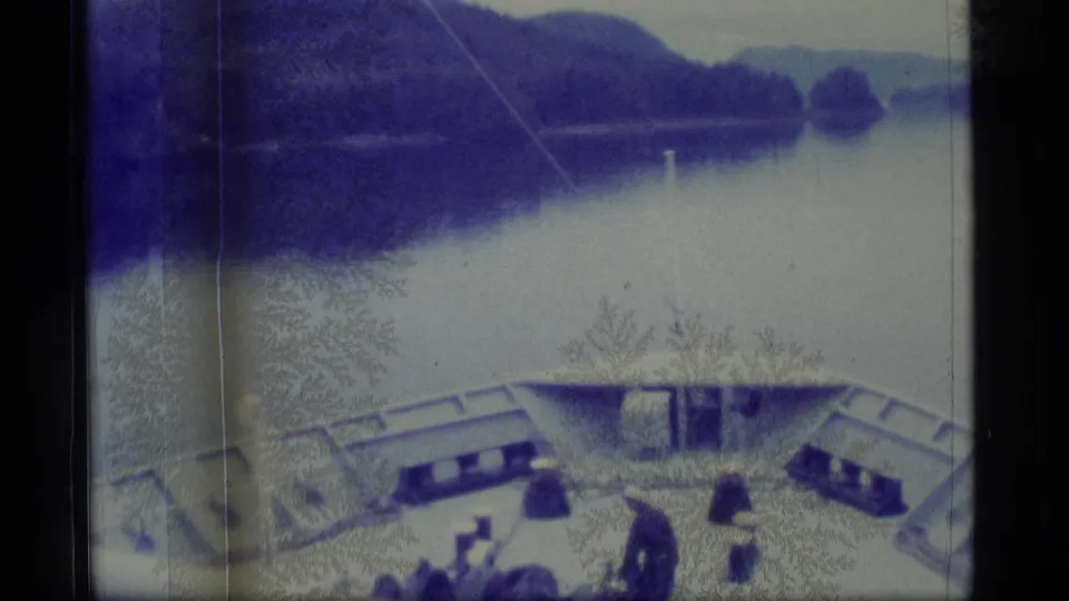1977 Alaska 8mm frame showing passengers and deck gear near the bow as the vessel moves along a mountain-lined waterway.