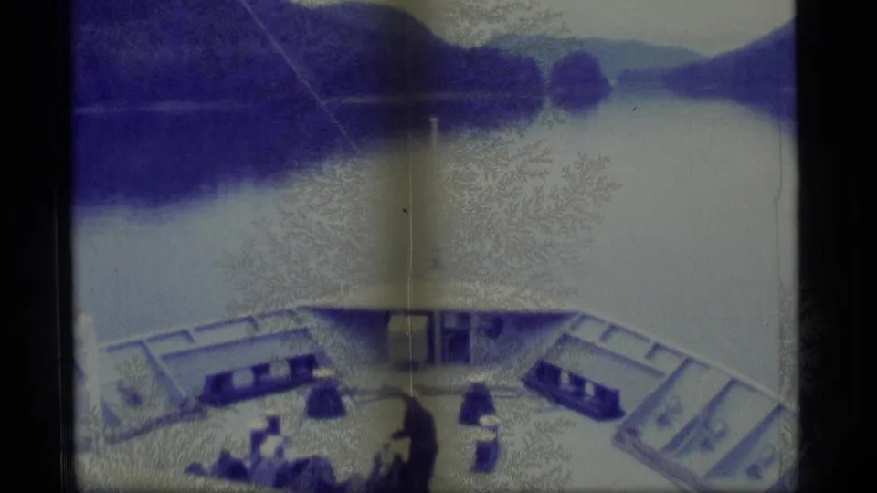 Blue-purple 1977 Alaska shipboard frame with passengers on deck, bow railings, and a quiet mountain waterway ahead.
