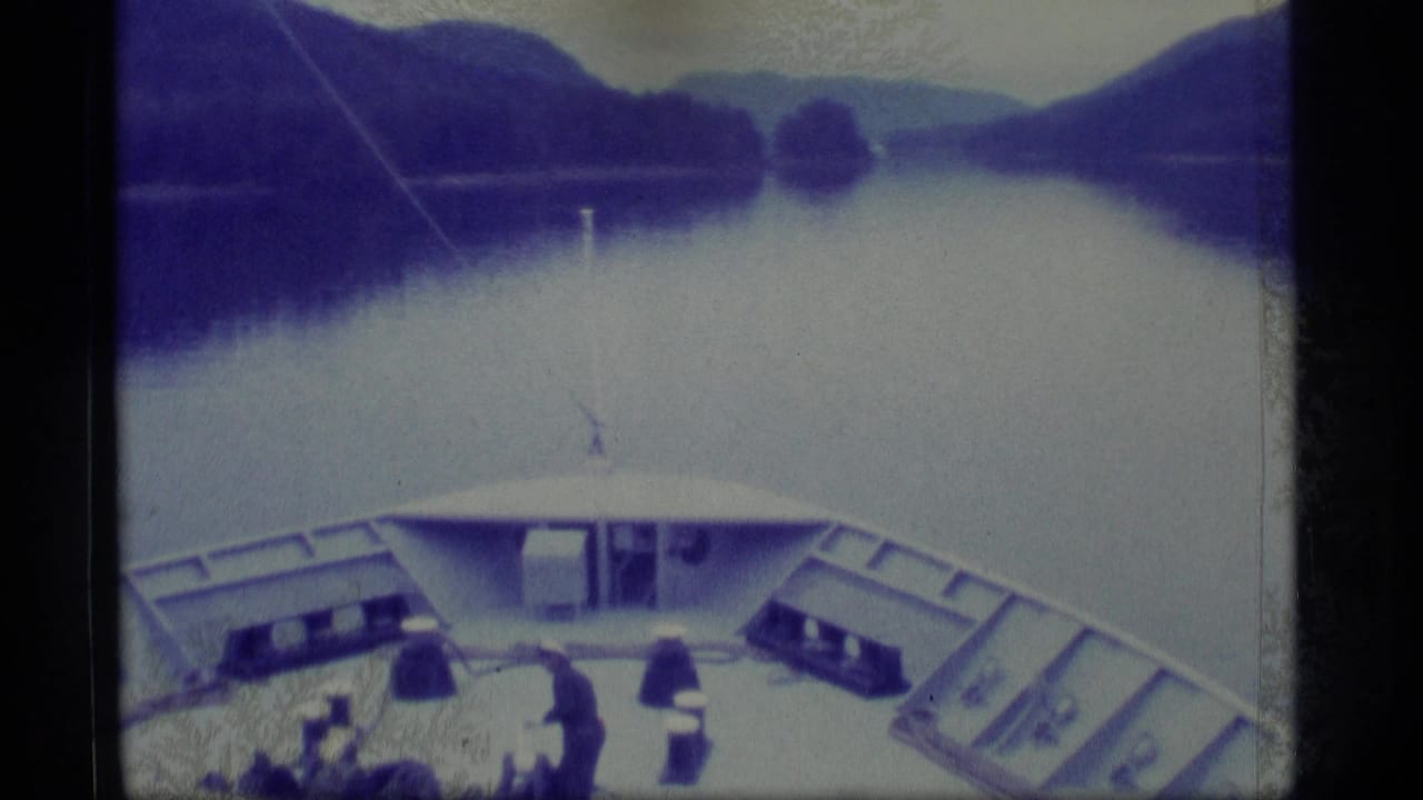 Bow of a vessel on calm water with mountains and a small standing figure on deck.