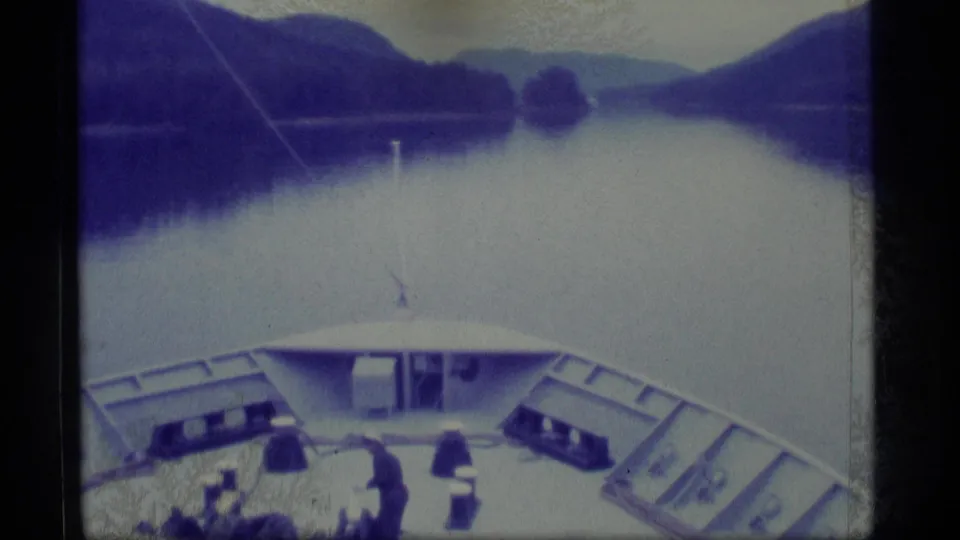 1977 Alaska 8mm frame looking over a vessel bow toward calm water, forested hills, and distant mountains.