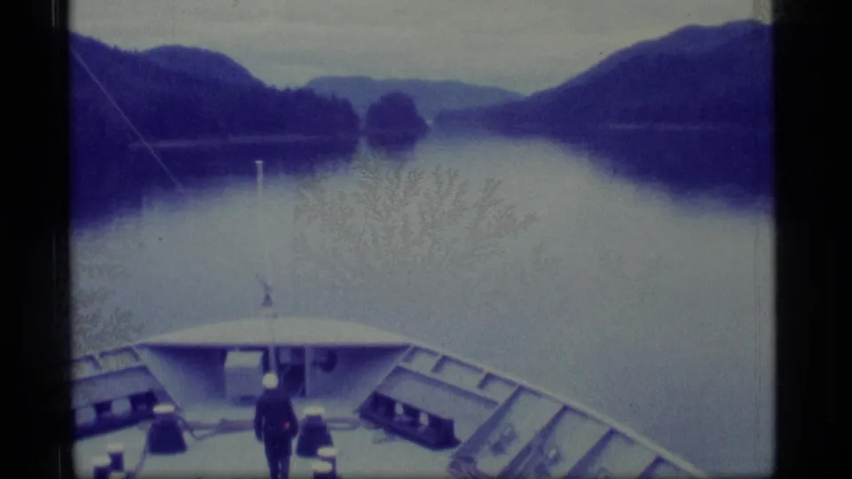 1977 Alaska home-movie frame with a deck passenger looking out over quiet water and forested mountain terrain.