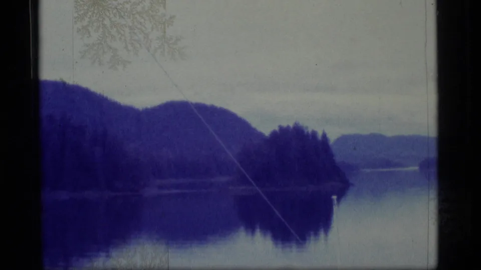 Alaska 8mm frame from 1977 showing a wooded shoreline, mountain slopes, and reflective water from a vessel viewpoint.