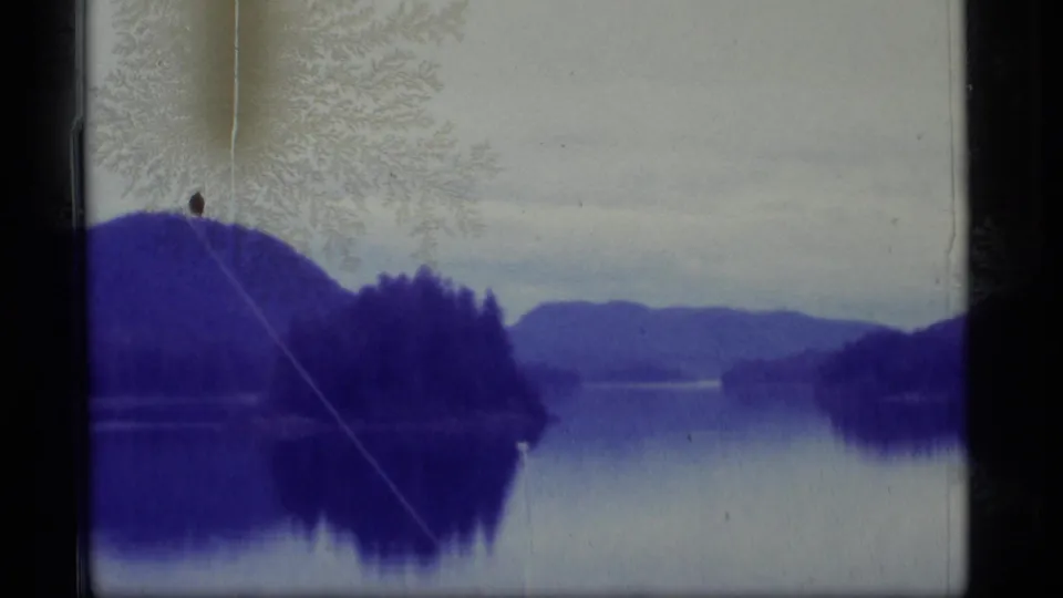 1977 Alaska 8mm view across a quiet lake toward forested hills and mountains, with visible scratches and aged-film texture.