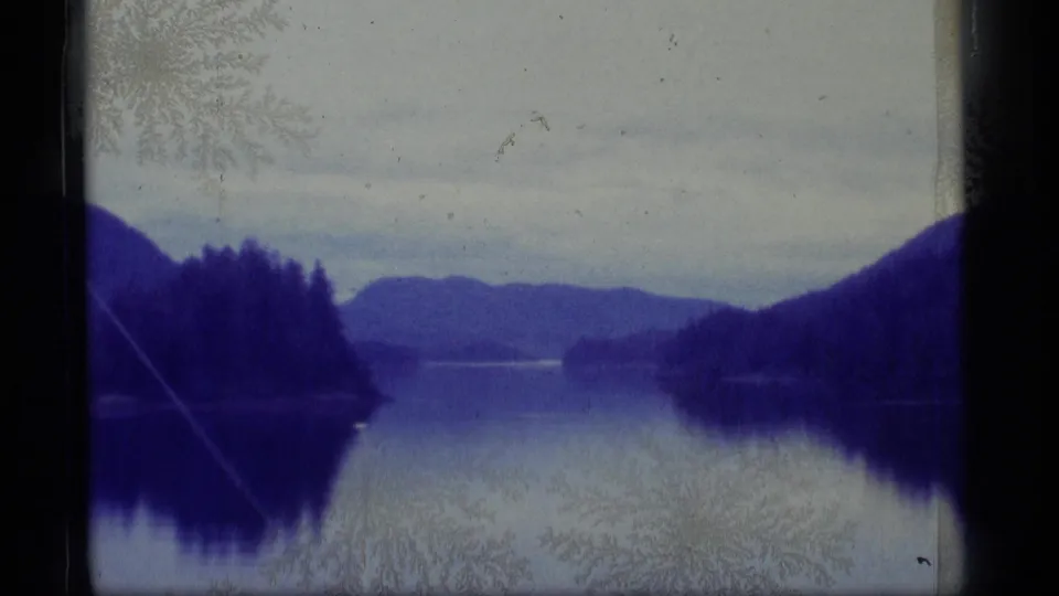 1977 Alaska home-movie frame showing a quiet lake between wooded hills, distant mountains, blue tint, and worn 8mm surface texture.