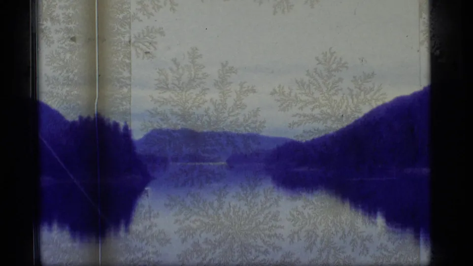1977 Alaska 8mm landscape frame of a misty lake, forested hills, reflected water, dark borders, and crystalline surface aging.