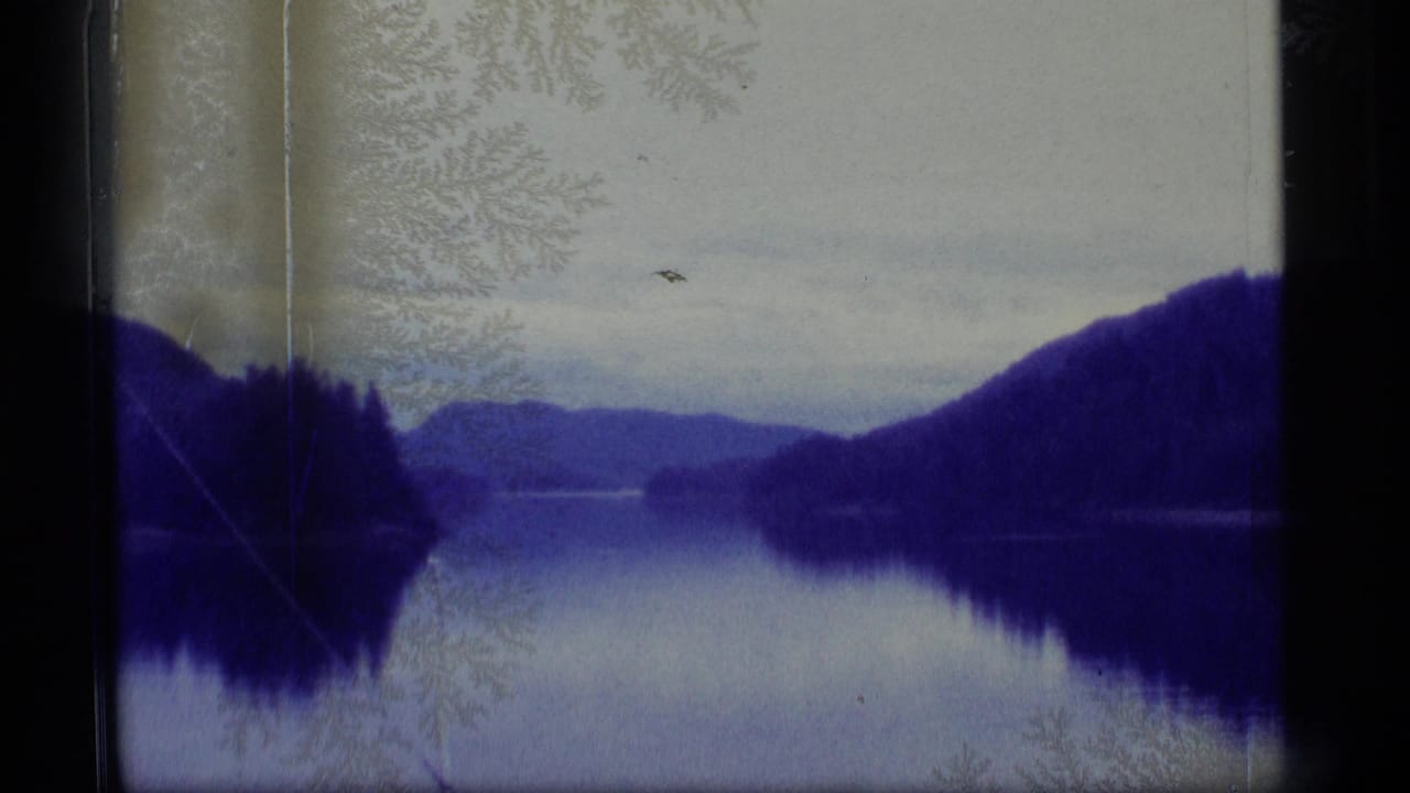 1977 Alaska 8mm frame showing a still lake reflecting dark forested hills and distant mountains beneath a cloudy sky.