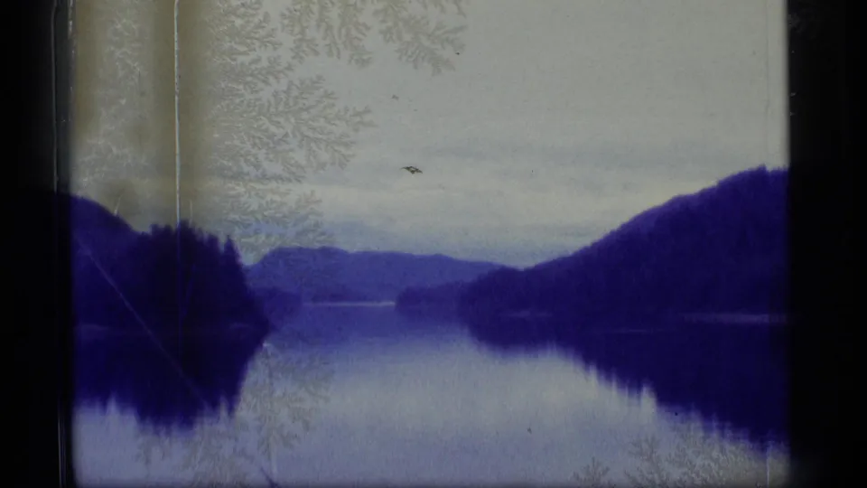 1977 Alaska 8mm frame showing a still lake reflecting dark forested hills and distant mountains beneath a cloudy sky.