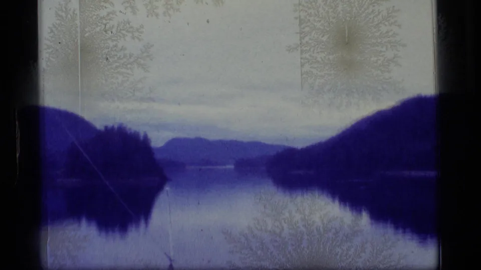 1977 Alaska 8mm frame of a quiet lake framed by dark hills and distant mountains, with gate edges and mottled film-surface texture.