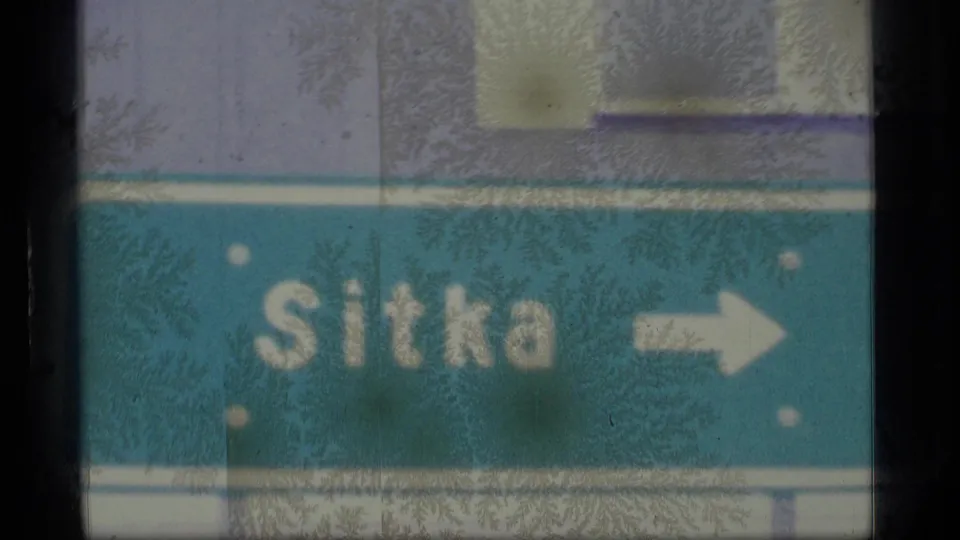 1977 Alaska 8mm frame showing a teal Sitka directional road sign with a white arrow, softened by film blur and surface marks.