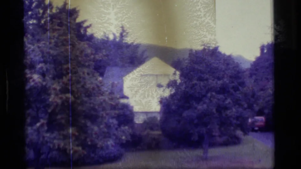 1977 Alaska home-movie frame with a white building screened by trees and a small red vehicle at the edge.