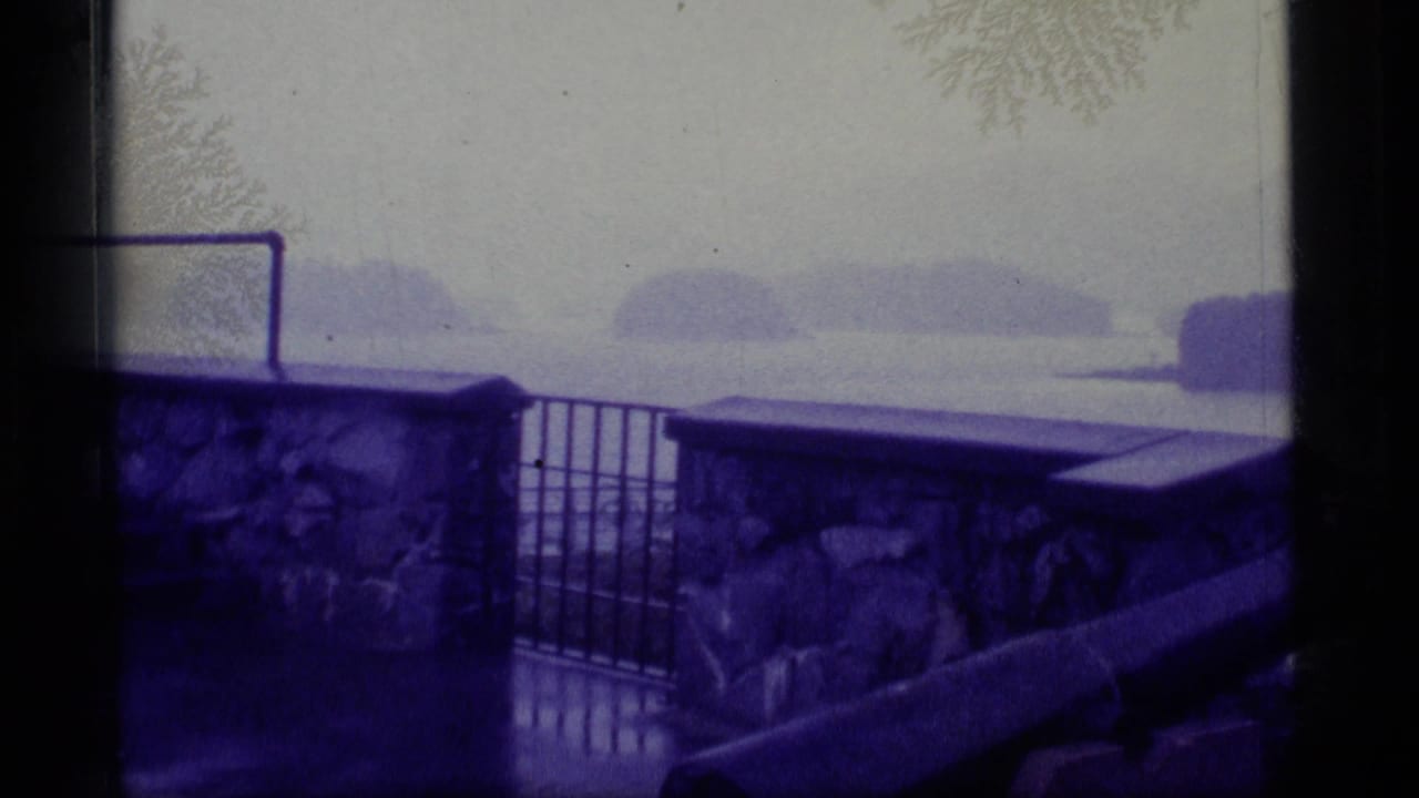 1977 Alaska 8mm frame showing a blue-purple tinted coastal view from behind a stone wall, gate, and foreground cannon.