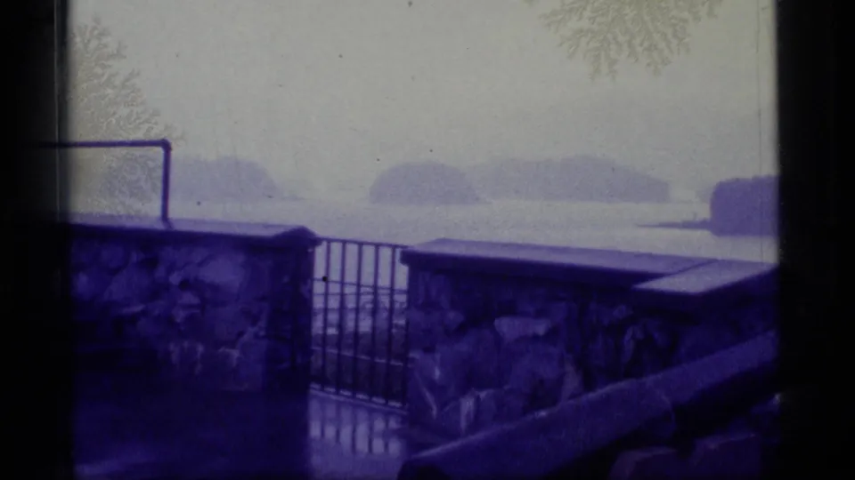 1977 Alaska 8mm frame showing a blue-purple tinted coastal view from behind a stone wall, gate, and foreground cannon.